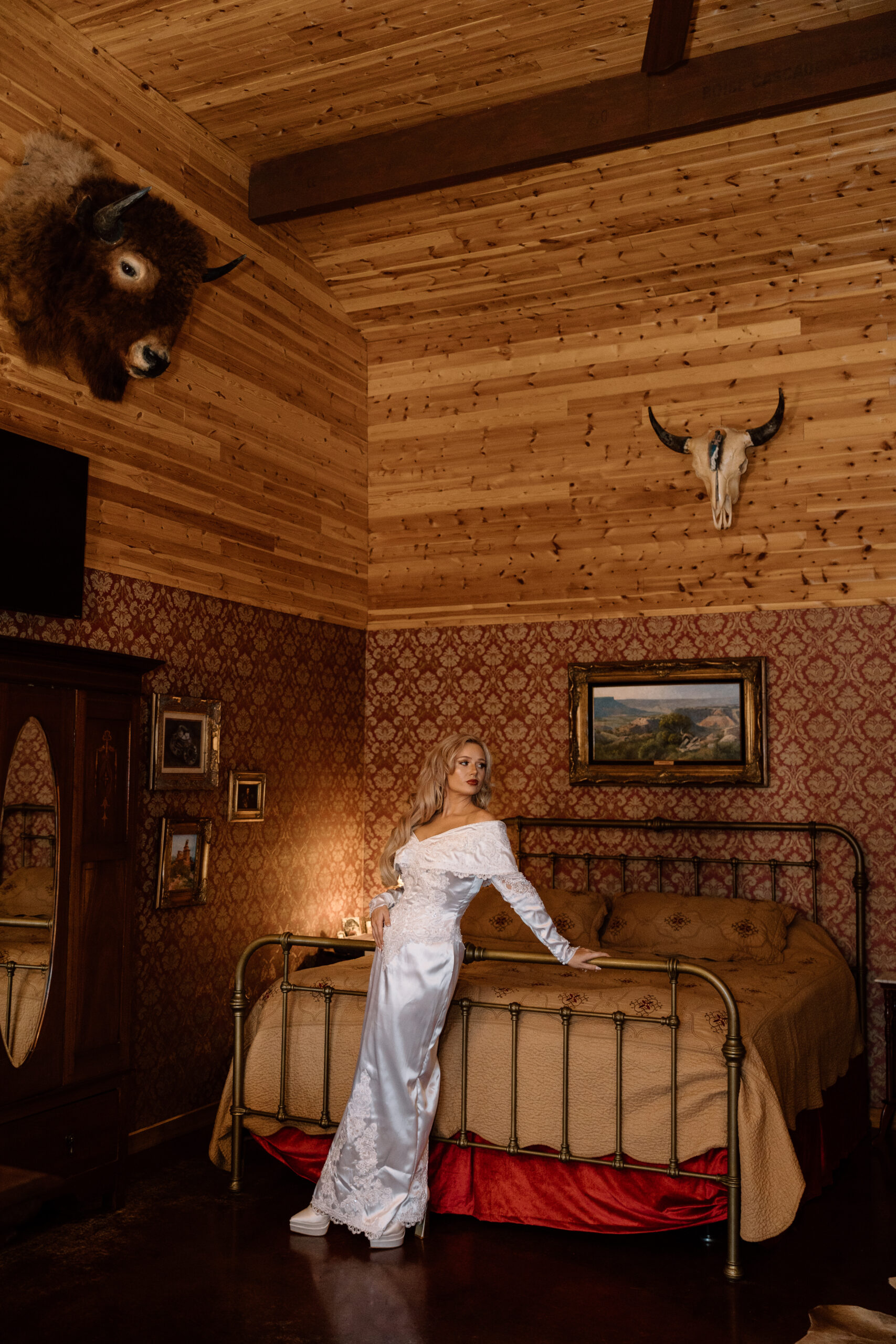 Bride in a vintage-style Western wedding dress leaning on a brass bed at The Hideout cabin near Palo Duro Canyon.