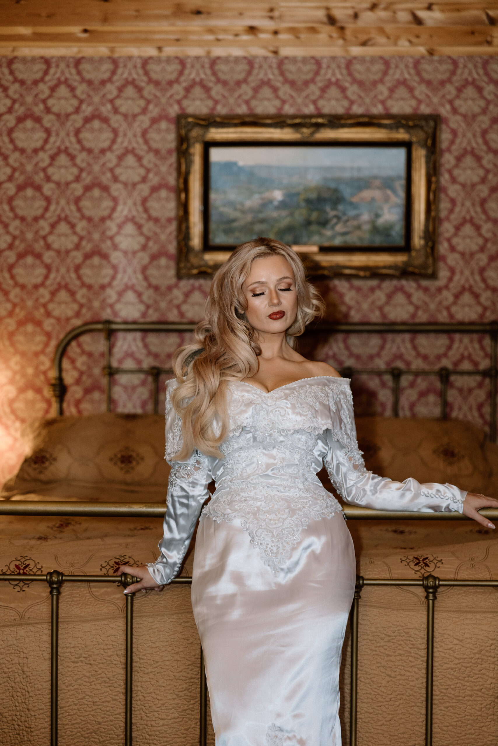 Bride in a vintage-style Western wedding dress leaning on a brass bed at The Hideout cabin near Palo Duro Canyon.